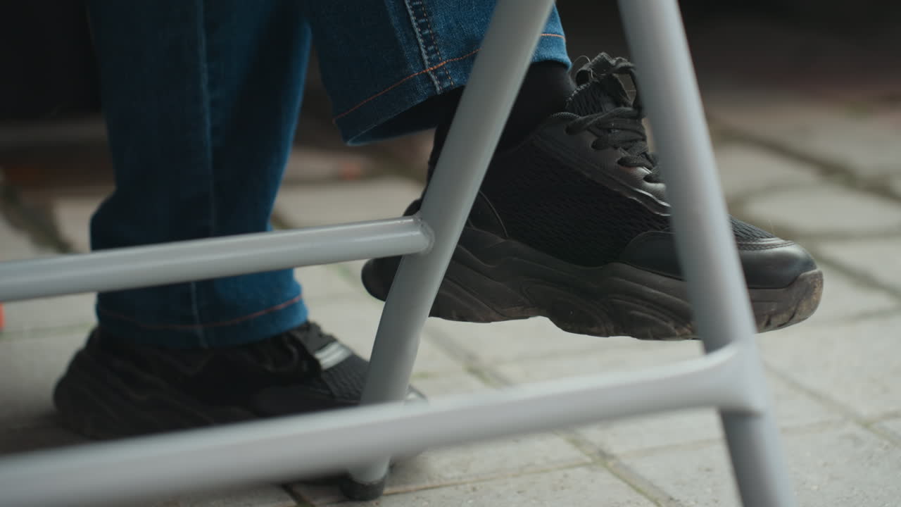 black sneaker under metal stool on pavement, closeup of leg in blue jeans resting on stool rung, outdoor cafe or sidewalk setting, casual commuter pausing midwalk, textured rubber sole and scuffed