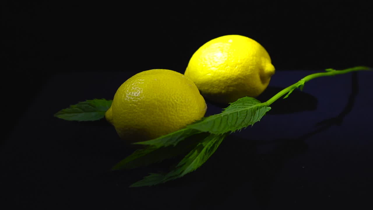 Close up view around yellow lemons and fresh mint sprigs stylized on a dark surface and black backdrop. Backlit highlights the texture of the fruits and herbs, peppermint stem casting dramatic shadow