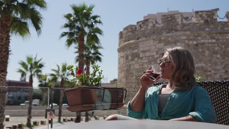 woman drinking iced coffee with palm trees and venetian tower of durres, kulla veneciane in the background in durres, albania. - closeup shot