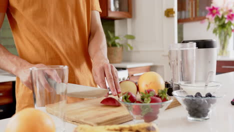 sección media de un hombre biracial cortando fruta para un batido saludable en la cocina, espacio de copia, cámara lenta