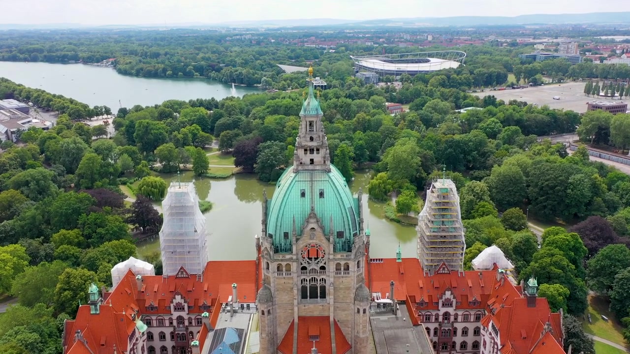 Immagini aeree mozzafiato che mostrano lo storico Nuovo Municipio di Hannover con la sua cupola iconica, affacciato sul pittoresco lago Maschsee e sul paesaggio verde circostante.