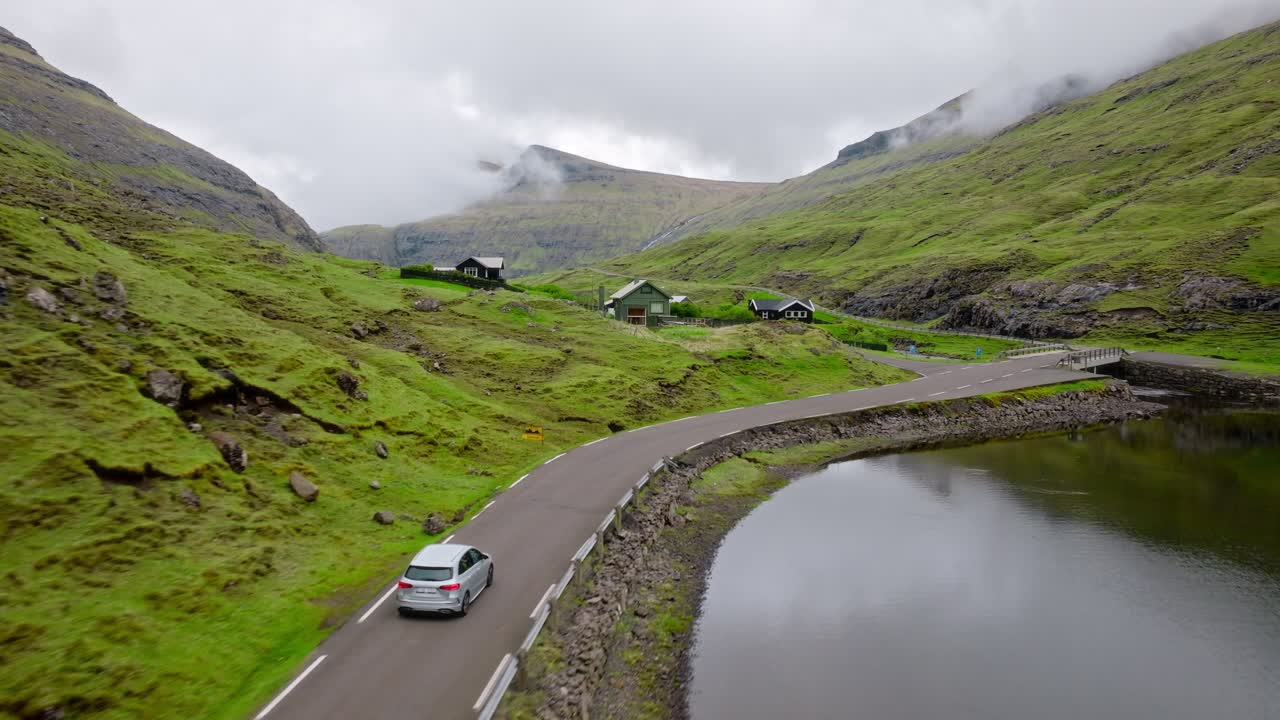A car drives through a scenic, foggy Faroe Islands valley with lush green hills