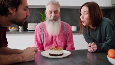 Un-Hombre-Feliz-Con-Cabello-Gris-Y-Una-Barba-Exuberante-Con-Una-Camisa-Rosa-Apaga-Una-Vela-En-Un-Pastel-De-Cumpleaños-Con-Sus-Hijos-Adultos,-Un-Chico-Moreno-Con-Barba-Incipiente-Y-Una-Niña-Con-Un-Suéter-Verde-En-Una-Cocina-Moderna.