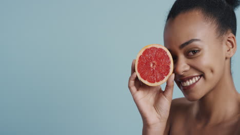 Porträt-Einer-Attraktiven-Jungen-Afroamerikanerin-Mit-Grapefruit,-Die-Lächelnd-Die-Natürliche,-Gesunde-Hautpflegeessenz-Genießt.-Schöne-Frau-Mit-Perfektem-Teint-Auf-Blauem-Hintergrund