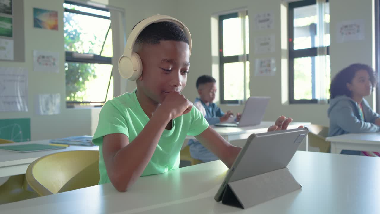 African teenage boy wearing beige headphones tapping tablet folio, following lesson at school desk