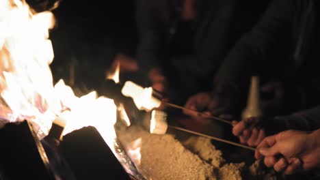 Closeup-view-of-hands-holding-sticks-with-marshmallows-and-frying-them-at-night.-Group-of-people-sitting-by-the-fire-late-at-night
