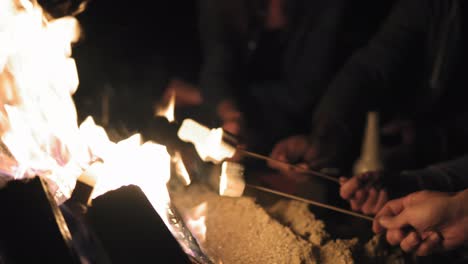 closeup view of hands holding sticks with marshmallows and frying them at night. group of people sitting by the fire late at night