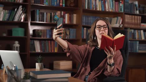 The-girl-is-sitting-in-a-chair-at-the-table,-holding-a-phone,-and-notebook,-or-book-in-her-hands