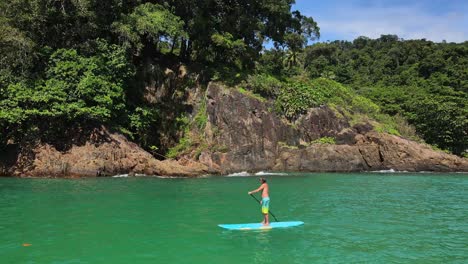 Toma-Aérea-De-Camiones-De-Un-Hombre-Caucásico-Haciendo-Ejercicio-En-Una-Tabla-De-Remo-En-Aguas-Turquesas-Tropicales-Claras,-Con-La-Costa-Tropical-De-La-Selva-En-Tailandia