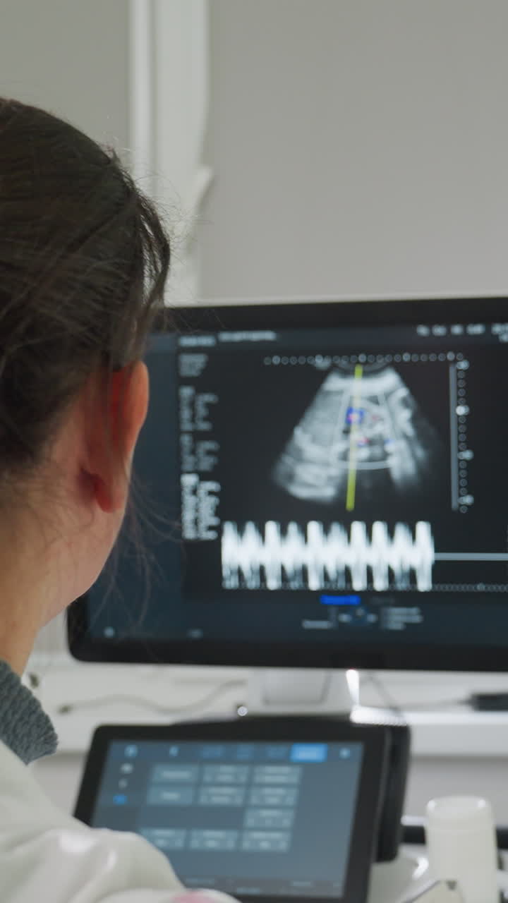 ecografista caucasico che esamina il monitor dell'ecografia fetale in una sala clinica silenziosa, tecnico che misura la forma d'onda del battito cardiaco sul display, radiologo che controlla le misurazioni biometriche sulla console, prenatale
