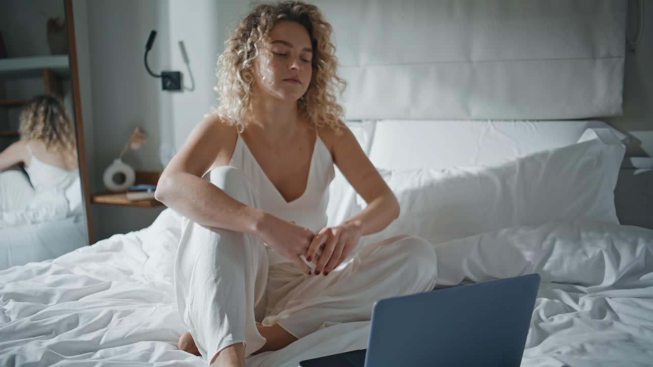 Relaxed girl using computer in cozy bed. Serene lady watching webinar at home