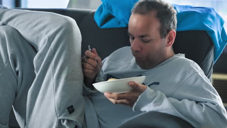 cinematic shot of white man with dark hair sitting relaxed on a black couch eating food while checking his phone in sweatpants in his apartment during a day