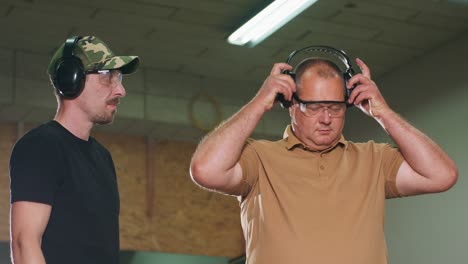 the trainee puts on protective glasses and earphones before taking a shot under the supervision of an instructor on a closed shooting range