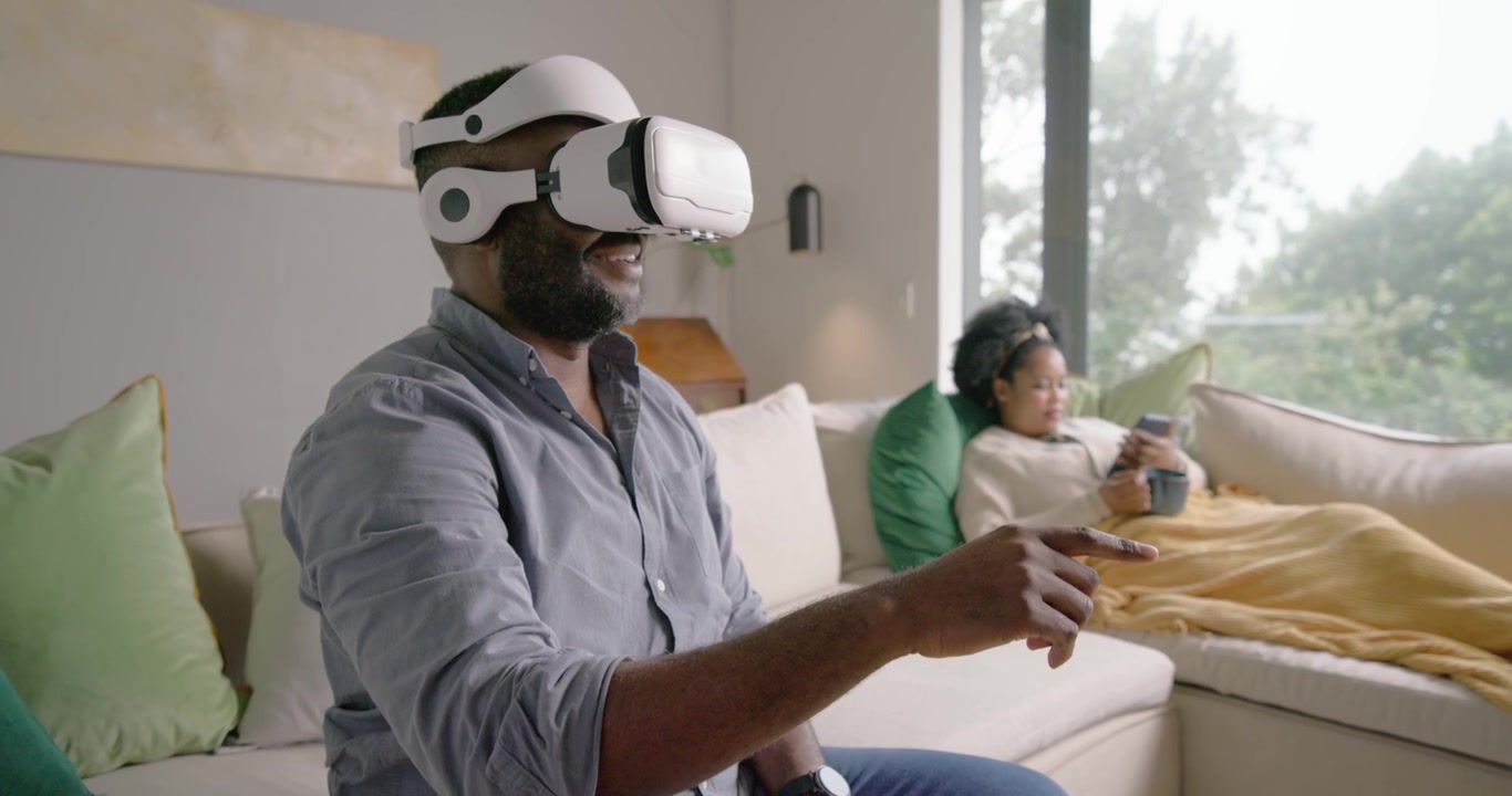 Coppia afroamericana sul divano, l'uomo che indossa un visore VR bianco e gioca in VR, la donna che scorre il telefono.