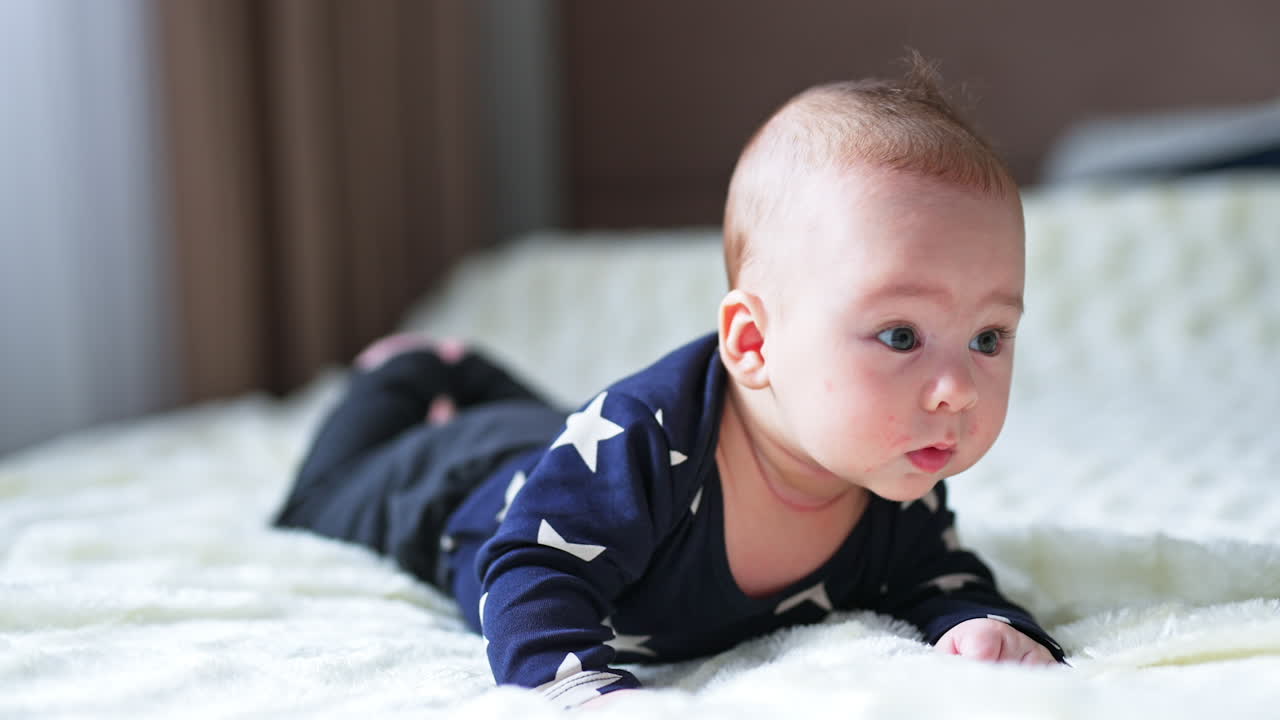 Little newborn baby looks puzzled gazing aside. Beautiful boy lies on belly keeping head up. Blurred backdrop.