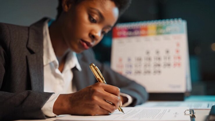 Focused Professional Writing: A Businesswoman Engaged in Task with Pen at Desk, Calendar in Background Representing Time Management and Productivity