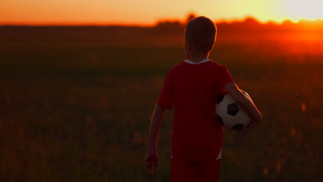 Der-Junge-Fußballspieler-Läuft-Mit-Dem-Ball-Auf-Das-Feld-Und-Träumt-Von-Einer-Fußballkarriere.-Bei-Sonnenuntergang-Blickt-Er-In-Die-Sonne.-Die-Kamera-Beobachtet-Den-Jungen,-Der-Bei-Sonnenuntergang-Mit-Einem-Fußball-über-Das-Feld-Läuft.