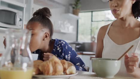 happy african american mother and daughter eating breakfast in kitchen, slow motion