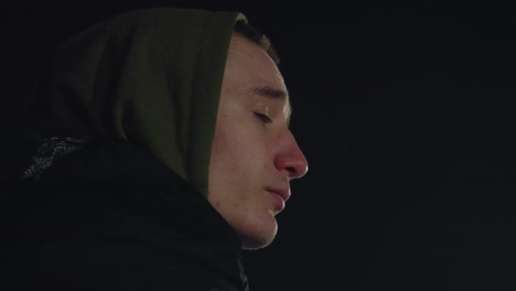 side profile of thoughtful boy wearing hoodie gazing into bright night light outdoors, with dark background suggesting quiet solitude and introspective mood in cold atmospheric scene