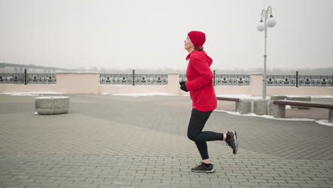 vista lateral de una mujer con capucha roja y gorra corriendo al aire libre durante el invierno en el pavimento entrelazado con elementos urbanos serenos como bancos, barandillas decorativas y alrededores nevados