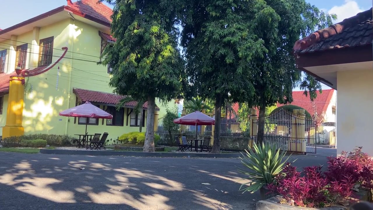 Halaman Cerah dengan Bangunan Kuning, Pohon Hijau, dan Tempat Duduk Luar Ruangan