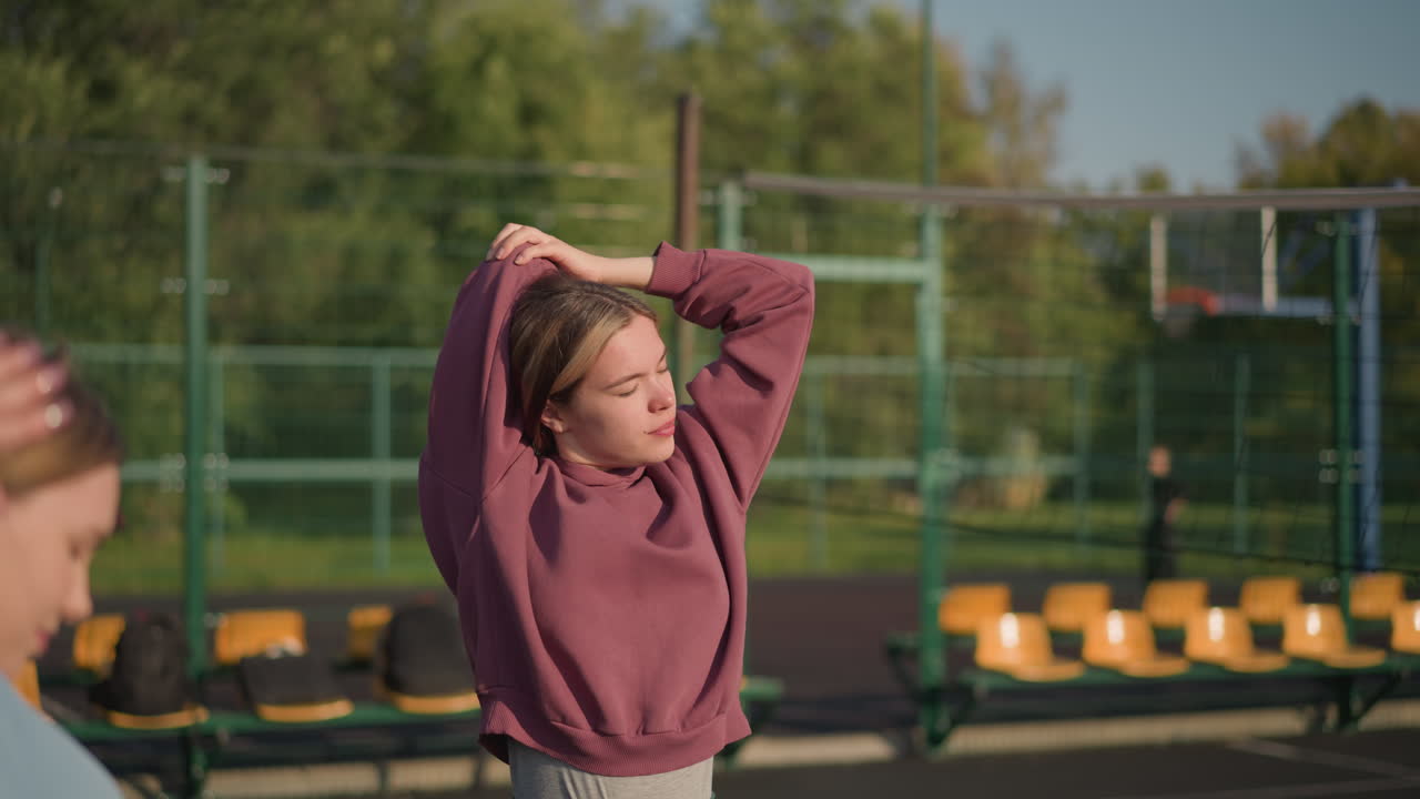 два молодых спортсмена тренируются на открытом воздухе с солнечным светом, освещающим их лица, на заднем плане расположены сиденья на стадионе, забор и размытый вид на кого-то, стоящего на расстоянии