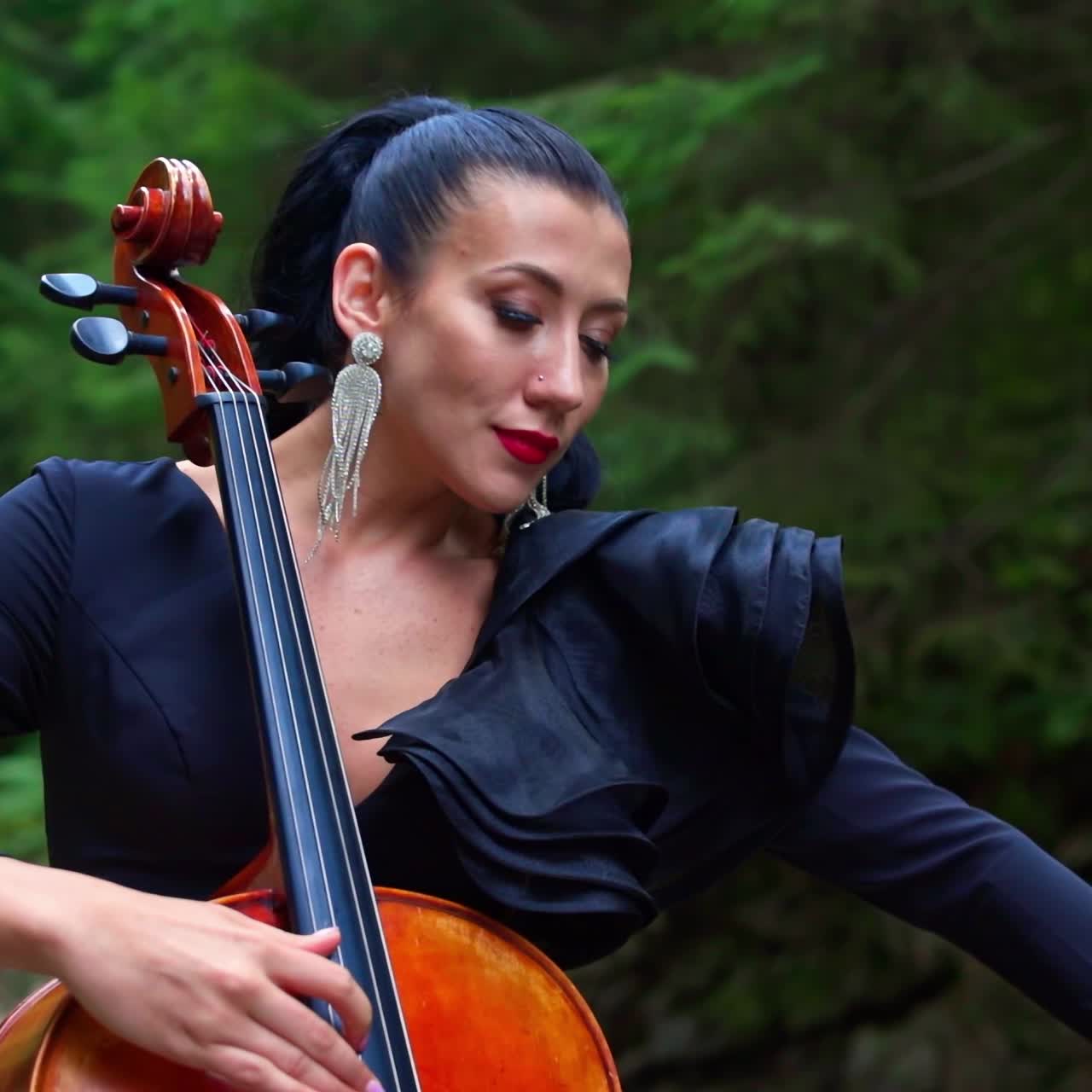 Attractive musician with cello. Beautiful woman in luxury black dress playing the musical instrument on summer nature background. Portrait of a female cellist.