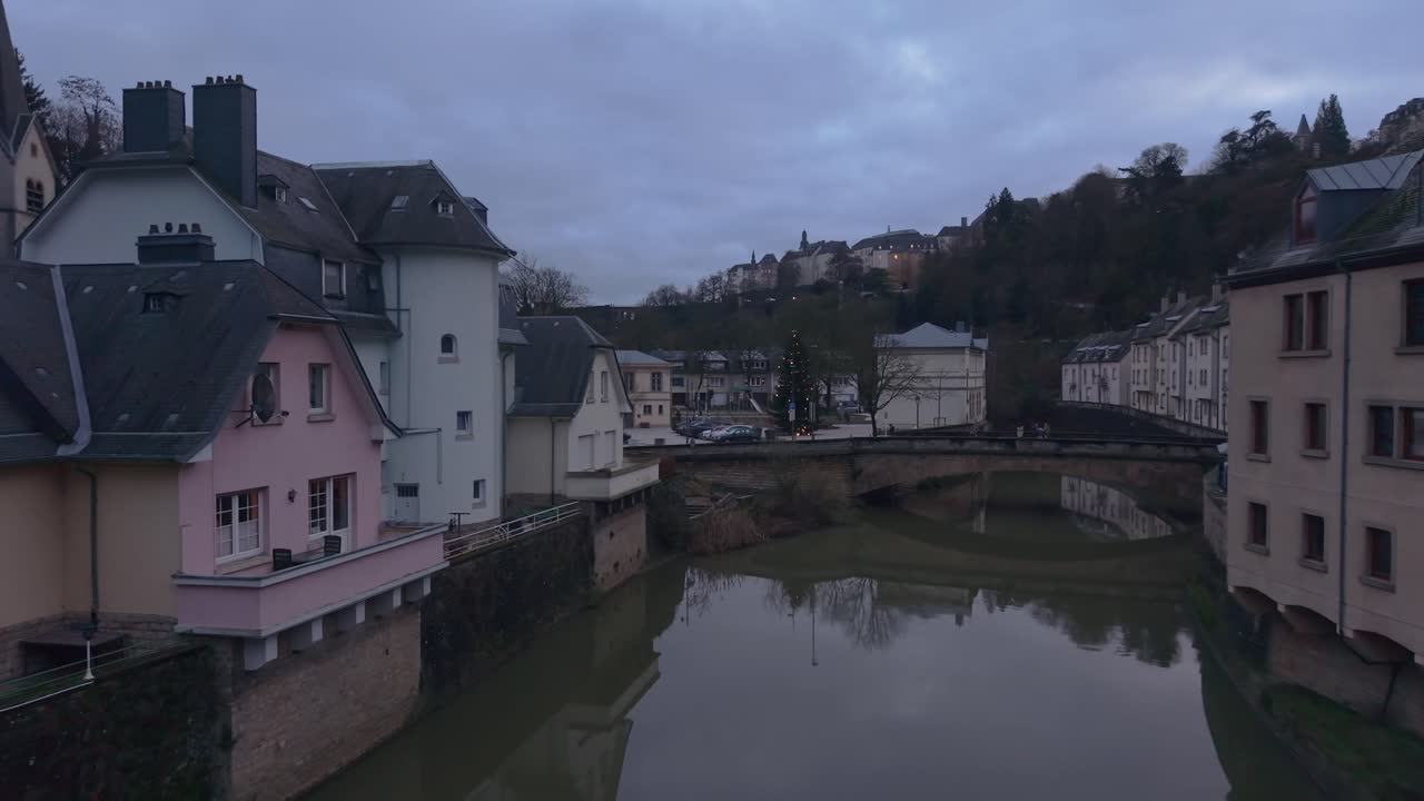 Lavtflyvende dronebevægelse over Alzette-floden i Luxembourg, der fanger maleriske huse langs floden, genspejlinger i vandet og det charmerende historiske bylandskab.