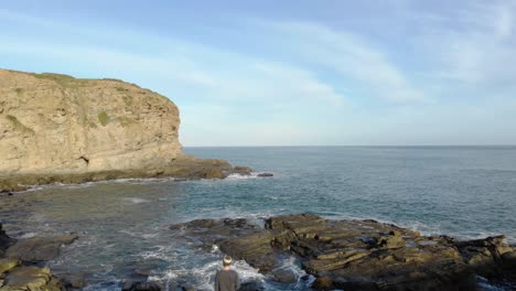 Aerial-shot-flying-over-a-man-standing-on-top-of-a-rocky-outcrop-on-the-edge-of-the-ocean-at-the-bottom-of-coastal-cliffs