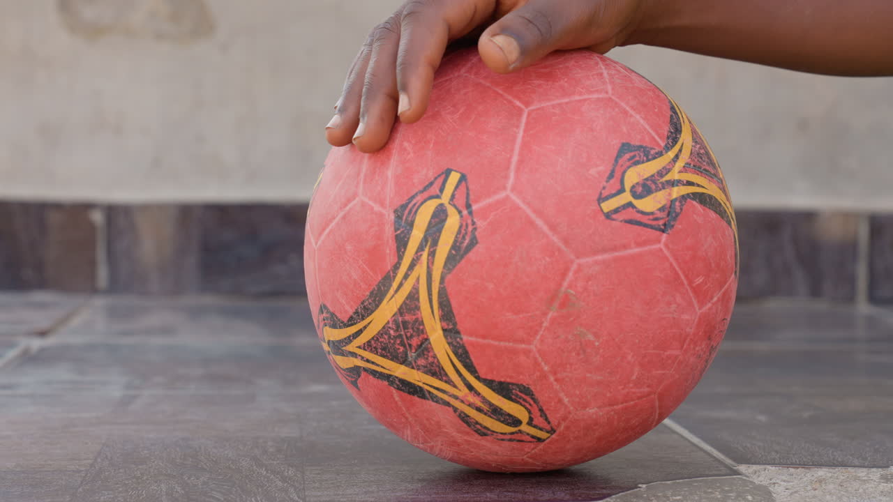Primo piano di una mano nera su un pallone da calcio rosso, che mostra la palla consumata e texturizzata su un pavimento in cemento piastrellato, la presa del giocatore con palmo e polpastrelli, con un'atmosfera da allenamento di strada urbano, focalizzato sui dettagli della superficie, ombra e polvere.