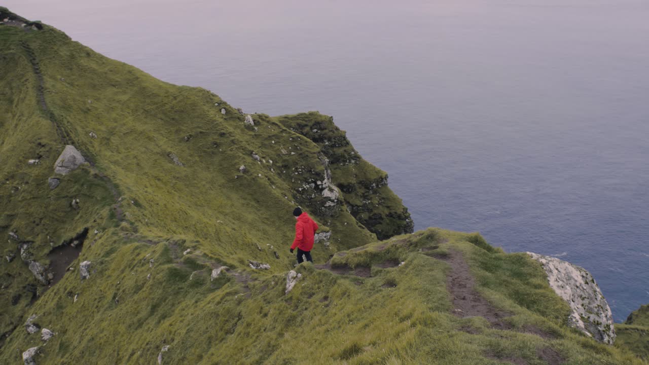 hombre con chaqueta roja camina y casi se desliza por un camino estrecho en un acantilado espectacular cerca del océano atlántico norte en el paisaje nórdico de kallur en las islas feroe