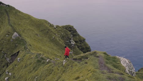 man in red jacket walks and almost slips on narrow path on spectacular cliff near the north atlantic ocean in the nordic landscape of kallur on the faroe islands