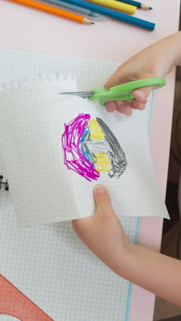 little child cuts perforated edge of sketchbook page with picture at desk upper view. toddler boy uses sharp tool to remove paper part at lesson