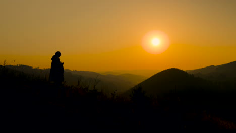 the silhouette of a woman watching the sunset and sitting down stump looking out at sunset with dark colors of mountains and hills in the background along with a orange sun in the background 4k 60fps