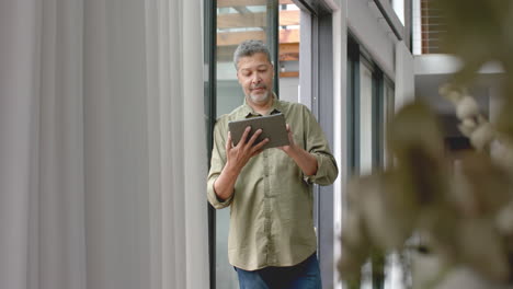 Glücklicher-älterer-Mann-Mit-Gemischter-Abstammung,-Der-Zu-Hause-Mit-Einem-Tablet-Am-Fenster-Steht,-Platz-Zum-Kopieren,-Zeitlupe