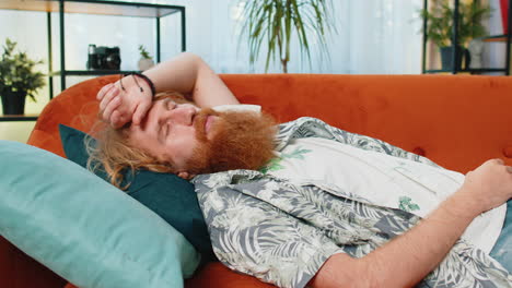tired caucasian redhead man lying down in bed taking a rest at home napping falling asleep on sofa