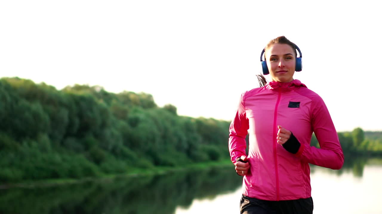 una chica con una chaqueta rosa y pantalones negros corre cerca del río con auriculares preparándose para el maratón