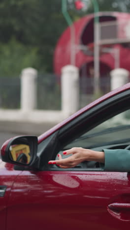 covered with raindrops red car moves past amusement park. positive female driver stretches hand through open window enjoying breeze while driving on blurred background