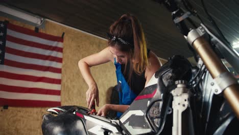 close-up shot: a girl auto mechanic repairs a motorcycle in her workshop with a screwdriver against the background of the us flag and tools in the garage