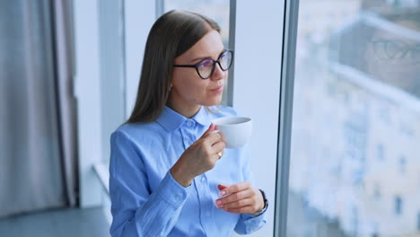 Young lady with long hair drinking coffee standing and looking into window. Relaxed calm female enjoying view from window.