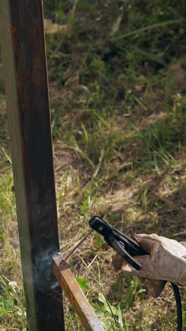 mature welder in work gloves uses powerful machine tool for fixing rusty carcass of metal fence in yard