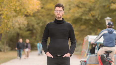 time lapse of a young caucasian man with a beard and glasses, dressed in a turtleneck sweater standing in an autumnal park, while passersby, people, and cyclists move and scurry around him