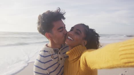 retrato de una feliz pareja hispana de pie en la playa, tomando una selfie