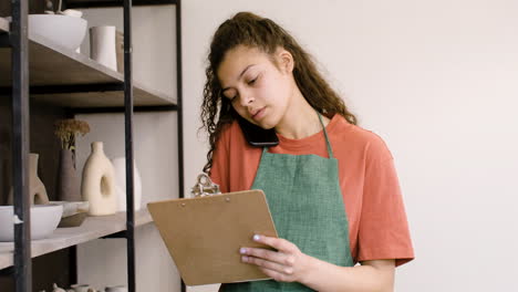 Young-Female-Clerk-Having-A-Call-And-Writing-On-A-Clipboard-In-The-Pottery-Shop-1