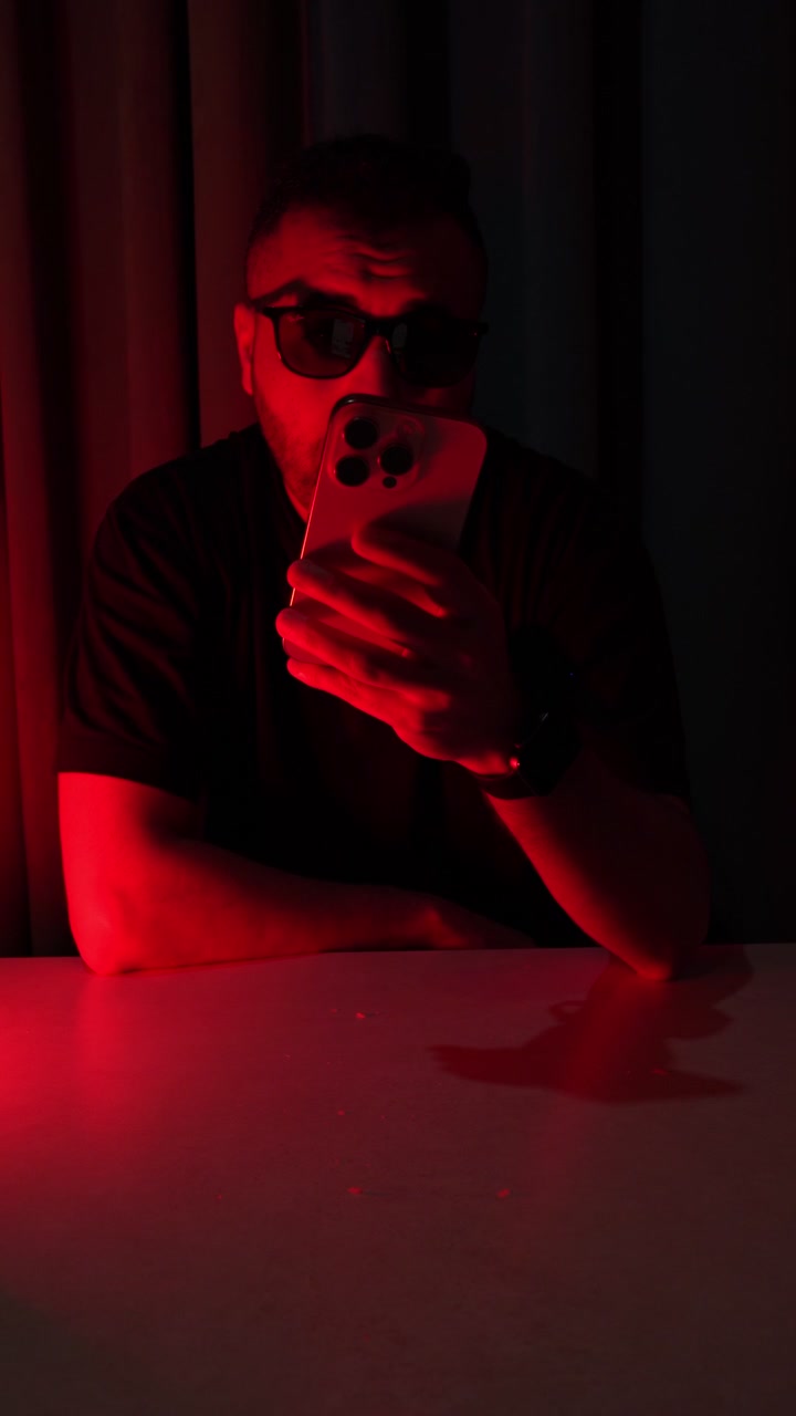 A man wearing sunglasses sits at a table in a dark room using a modern smartphone. Dramatic red studio lighting illuminates his face and the mobile device during this intense indoor scene.