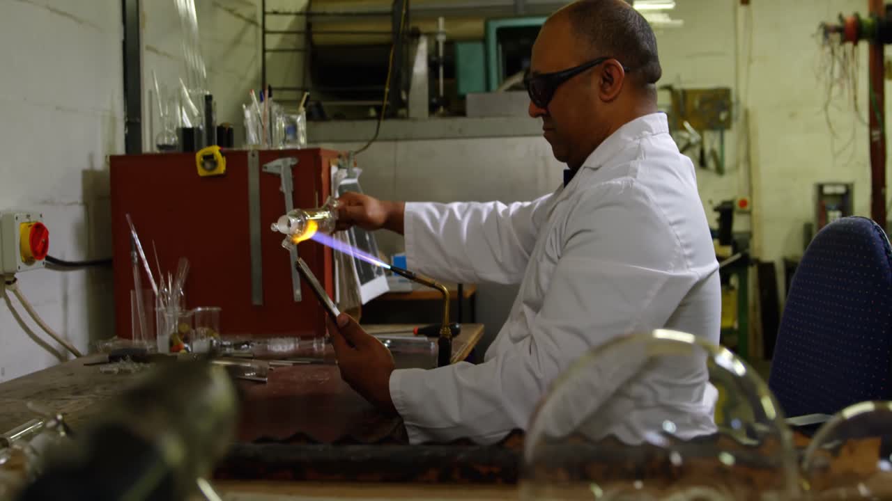 vista lateral de un trabajador masculino maduro trabajando en una tableta digital mientras sopla vidrio en una fábrica de vidrio 4k