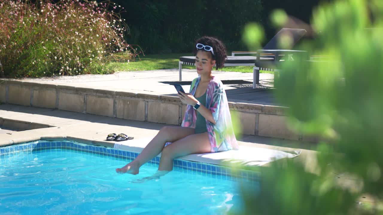 Adult female holding device at pool edge with sunglasses, tapping screen and relaxing, feet in pool