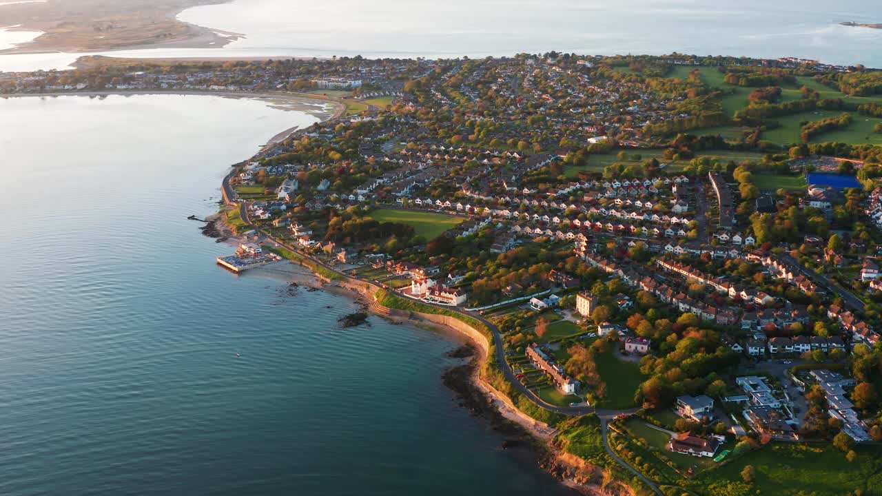 Aerial orbit of Sutton South coastline during golden hour, with homes, calm sea, and a scenic road view