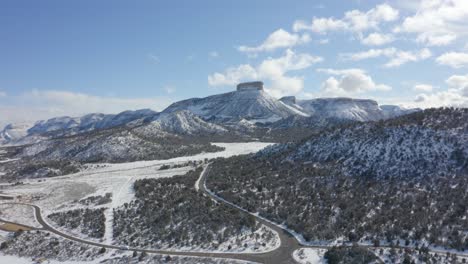 An-aerial-view-of-a-road-passing-through-snow-capped-mountains-with-a-razor-sharp-peak-clothed-in-green-flora-during-a-bright-day-is-surrounded-by-clouds