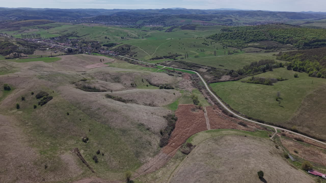 Vista aerea di dolci colline verdi e una valle con una strada tortuosa che porta a un villaggio lontano sotto un cielo sereno